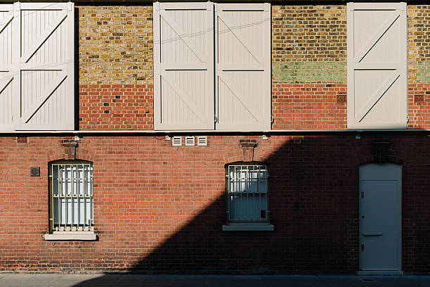 A weathered brick building with a row of barred windows, casting long shadows on the sidewalk. The facade shows signs of aging, with crumbling bricks and peeling paint, evoking a sense of history and decay. The interplay of light and shadow adds depth and contrast to the urban scene.