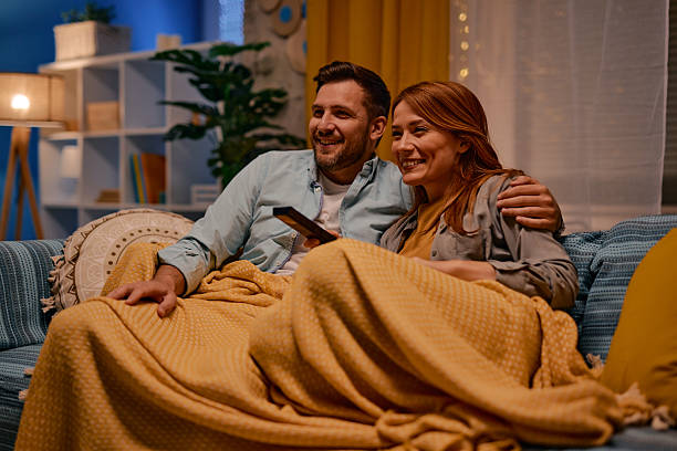Happy couple watching television covered by blanket on sofa at home