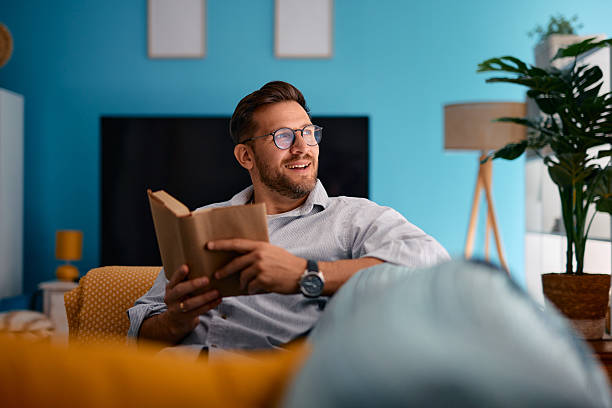 istock Man smiling and holding a book while sitting on a sofa at home 2217441009