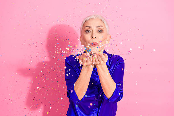 istock Confident modern senior woman joyfully blowing confetti in studio against pink background expressing celebration and vibrant lifestyle 2217441748