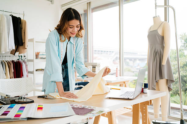istock Young woman fashion designer working in a modern studio, selecting fabric while referencing designs on a laptop. Small business owner creating custom clothing for online or boutique retail markets. 2217448905