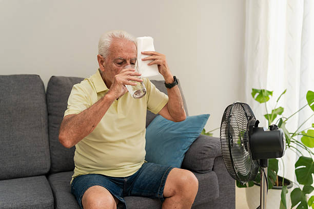 istock Senior man refreshing drinking water and wiping sweat with a towel sitting on sofa with electric fan 2217450181