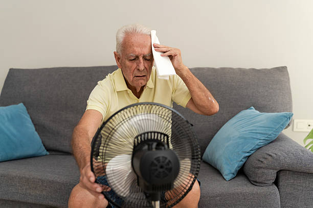 istock Elderly man cooling off with electric fan and wiping sweat from forehead while sitting on sofa 2217450201