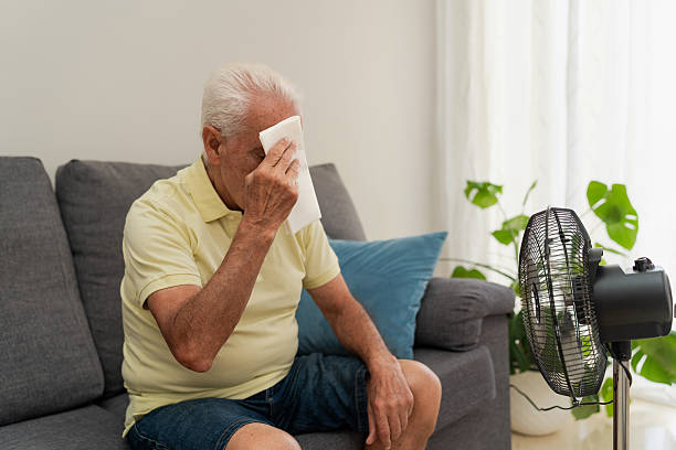 istock Elderly man wiping sweat from his forehead while sitting on sofa at home using electric fan 2217450205