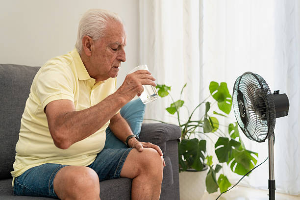 istock Senior man drinking water and cooling with fan during hot weather while sitting on sofa at home 2217450221