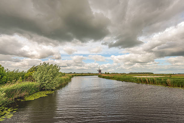 A characteristic Dutch polder landscape featuring a historic thatched hollow post windmill, set beside a wide canal. Captured on a windy spring day under a dramatic sky filled with dark clouds