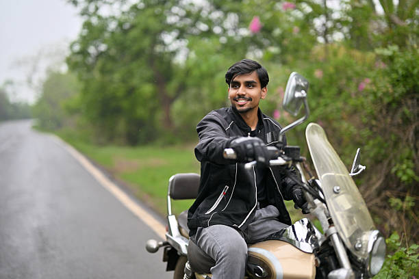 Happy young man in a black riding jacket and gloves enjoying a motorcycle ride on a peaceful rural road surrounded by greenery. The clear windshield and protective gear reflect a sense of adventure, safety, and freedom. The image captures the spirit of road travel, outdoor lifestyle, and solo exploration.