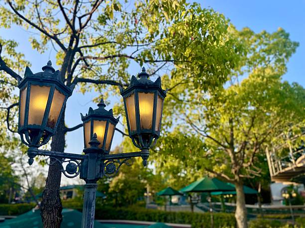 Three ornate, vintage-style street lamps are illuminated against a backdrop of lush green trees and a clear blue sky