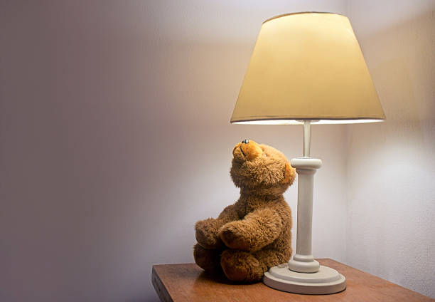 Brown teddy bear sitting on a wooden nightstand, gazing up at a softly glowing lamp, creating a warm and comforting bedtime atmosphere perfect for relaxation and sweet dreams