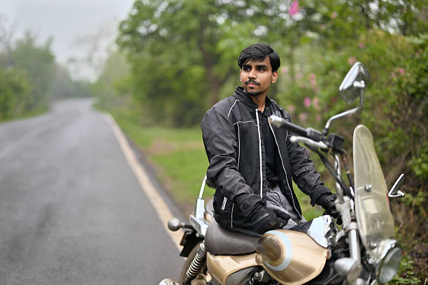 Confident Indian man wearing a black biker jacket and gloves, seated on a classic motorcycle parked on a quiet countryside road. Surrounded by lush greenery and soft natural light, it captures the spirit of freedom, travel, and self-discovery. The scene reflects adventure, solo road trips, and a peaceful connection with nature.