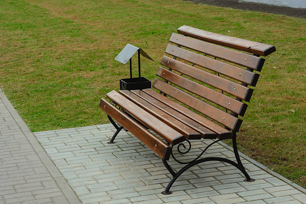 Wooden park bench with a trash can nearby, placed on a paved path beside a modern building with large windows. Peaceful urban setting for outdoor relaxation