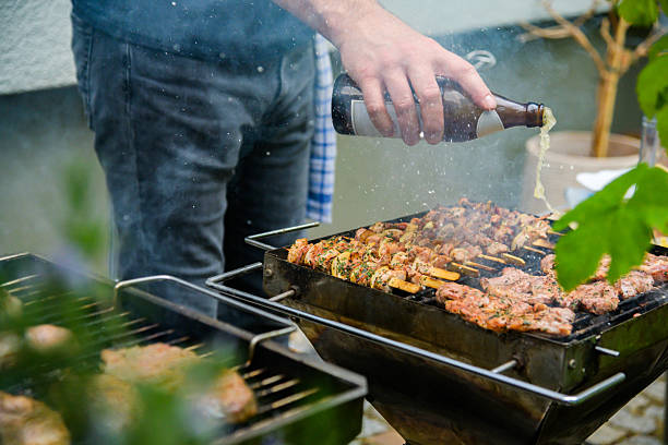 A person grills an assortment of meat skewers on a barbecue, enhancing flavors by pouring beer from a bottle. The outdoor environment creates a casual and inviting summer cooking atmosphere.