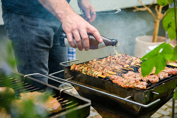 A person grills an assortment of meat skewers on a barbecue, enhancing flavors by pouring beer from a bottle. The outdoor environment creates a casual and inviting summer cooking atmosphere.
