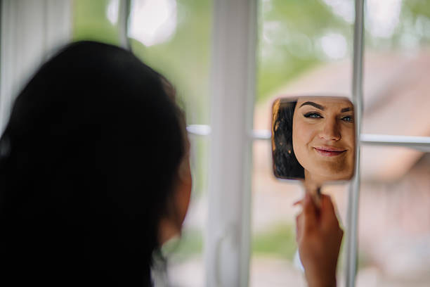 istock A smiling woman with elegant makeup admires her reflection in a handheld mirror by the window, surrounded by soft daylight and natural tones. 2217496178