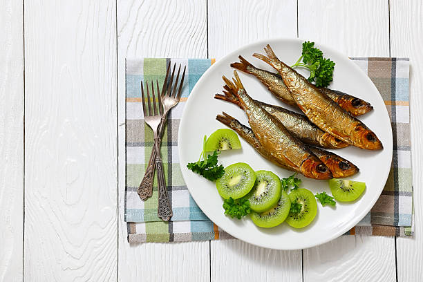 Hot smoked Baltic herrings with kiwi fruit slices and parsley on a plate on white wood table with forks, close-up, flat lay, free space, real photo, not AI generated