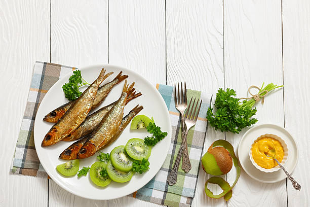 Hot smoked Baltic herrings with kiwi fruit slices and parsley on a plate on white wood table with forks, horizontal view from above, flat lay, free space, not AI generated