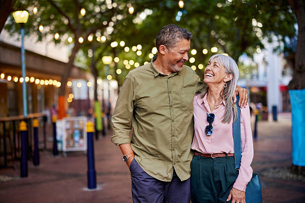 Happy multiethnic man and smiling  woman enjoy a romantic evening outdoors. Mature couple shares a loving moment while walking in a european city. Smiling senior couple walks closely together under string lights with copy space.