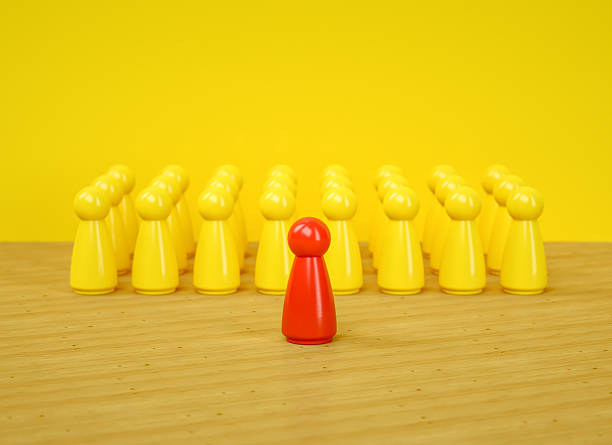 A compelling visual metaphor for Leadership, Individuality, and Standing Out From The Crowd. A single, distinct red game pawn stands confidently before a large, uniform group of yellow pawns, representing a Leader addressing their Team, a CEO presenting to their company, or an influencer speaking to an audience. The composition on a wooden surface with a Yellow Background powerfully illustrates concepts like Authority, Management, public speaking, and having a unique Vision. This image is perfect for business presentations, corporate training materials, and articles on personal Development, Confidence, and the courage to be different.