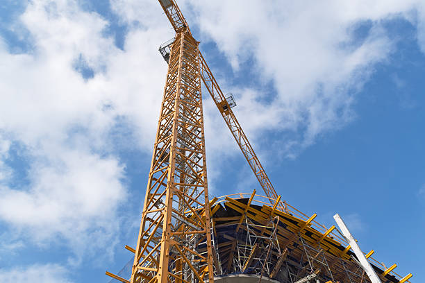 A towering yellow construction crane rises confidently above an unfinished building, reaching into the vivid blue sky scattered with clouds, symbolizing growth and ambition.