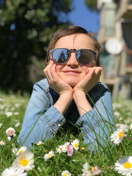 Child is lying in a field of daisies, wearing sunglasses and a denim jacket, with a relaxed and happy expression. The sunlight enhances the vibrant green grass and white flowers, capturing a serene, joyful moment in nature