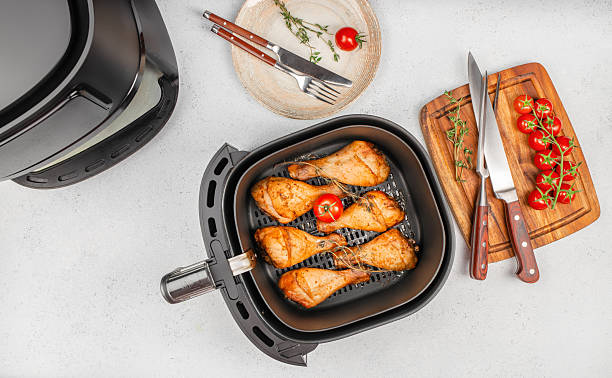 Grilled chicken drumsticks in air fryer basket with tomatoes, herbs and kitchen tools on countertop. top view. copy space.