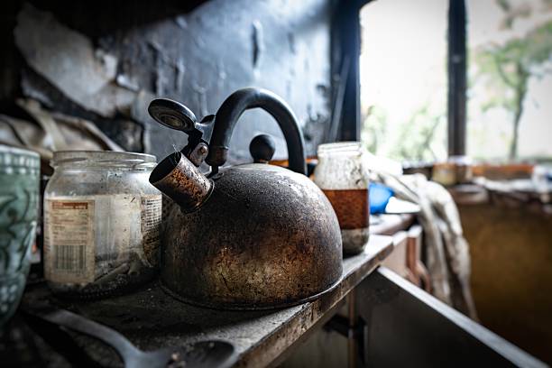 A dirty kettle sits on a kitchen table covered in soot and grime, reflecting the neglected and abandoned state of the house.