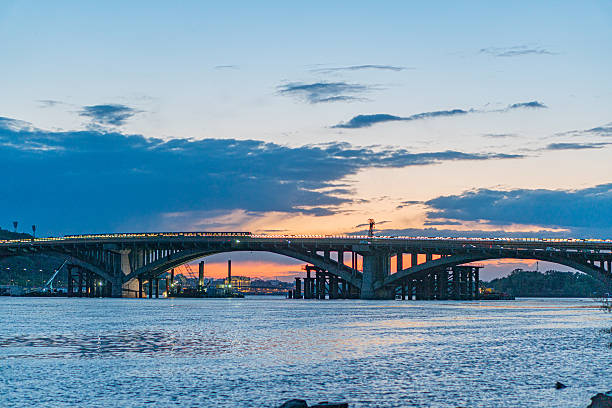 istock Dusk scene at Kievs Dnepr River bridge Long suspension bridge with multiple spans, reflecting on calm water Illuminated urban structures along the bridge Warm orange horizon merging with lighter 2217529812