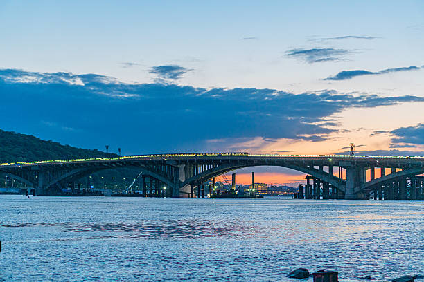 istock Suspension bridge over Dnepr River at sunset, calm dark blue harbor, multiple white pillars, glowing lights, serene scene, mixed sky tones deep blues, warm oranges, reflecting colors on water, smal 2217529822