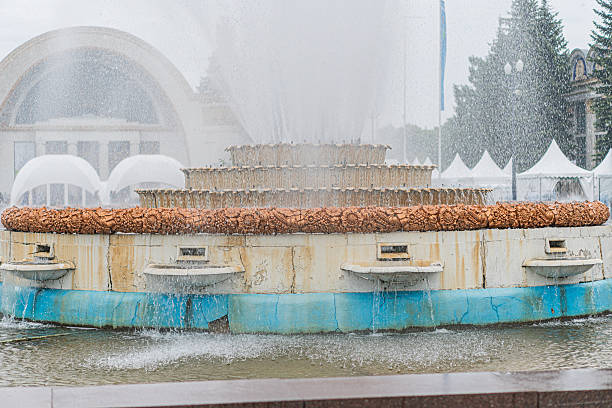 Large circular fountain in a public space, operating with multiple water jets The structure has varying tiers and flower-like elements, contrasting against light-toned pavement Clear sky with fluff