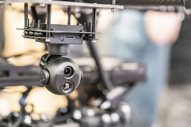 A combat quadcopter drone is shown, attached to a robotic arm in operation within an industrial workspace The arm moves from middle left to top right, with a spherical camera assembly at its end cap