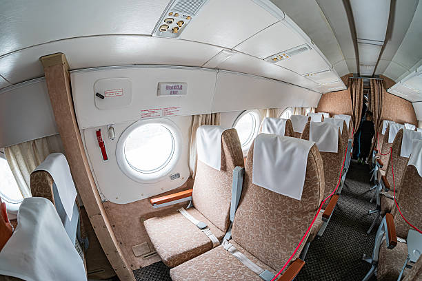 istock Small airplane cabin interior, three-seat rows covered in white sheets, overhead bins empty, neutral colored walls and carpeted floor, soft ambient lighting, patterned carpeted aisle, partial view of 2217532645
