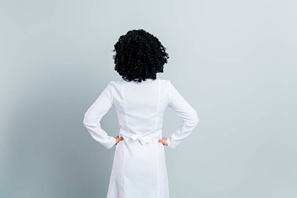 A rear-facing female medic, wearing a clinical white lab coat, stands confidently against a plain gray background, emphasizing dedication.