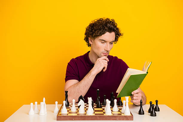 istock Young man with curly hair reading a book while sitting at a table with chess pieces on a yellow background 2217539033