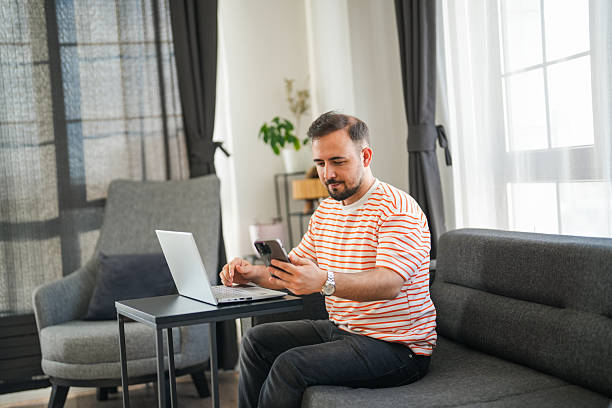 istock Man sitting on sofa in living room working on laptop while also looking at his phone 2217546874