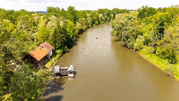 A charming old mill with a rusted roof sits on the bank of a calm river, surrounded by lush green trees under a bright daytime sky.