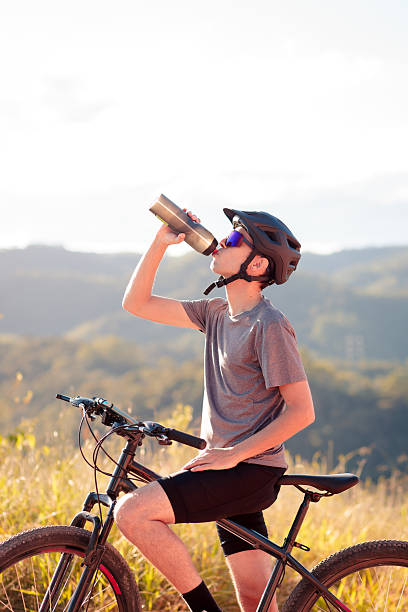 A man cyclist wearing a helmet and bright yellow jersey takes a refreshing drink from a blue water bottle while riding along a rocky, nature-filled trail.