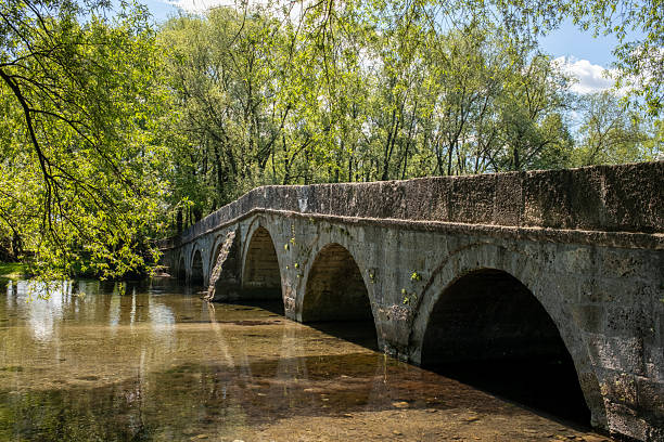 istock Historic stone bridge spanning a shallow river with lush green trees on a sunny day Arched architecture in a natural landscape 2217559217