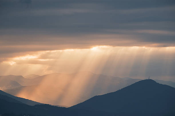 Sun rays pierce through overcast clouds, illuminating a series of mountains. A tranquil, scenic view evokes a sense of peace and the beauty of nature.