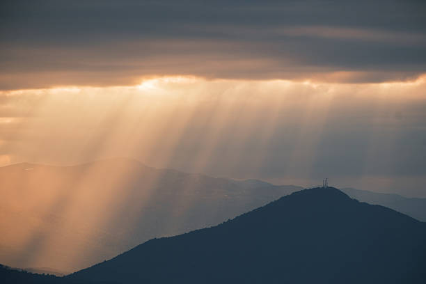 Golden sunrays stream through dark clouds, highlighting the misty ridges of a mountain range. A tranquil and captivating landscape scene at twilight.