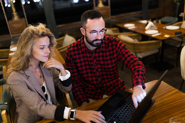 istock Colleagues collaborating in a cozy restaurant setting, focusing intently on a laptop screen for a project discussion. Woman thoughtfully observing while man actively points at the display, enhancing teamwork and creativity. 2217561838