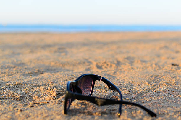 Sunglasses lie on a sandy beach with ocean backdrop. Symbolizes relaxation, vacation, summer vibes, and carefree lifestyle. Ideal for travel or leisure themes.