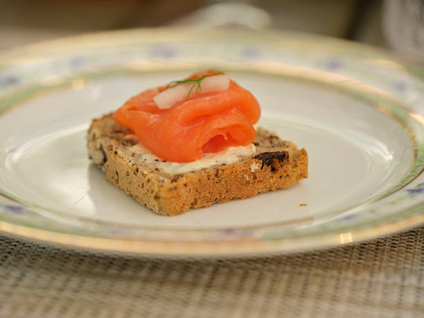 A visually appealing close-up of an appetizer with smoked salmon elegantly placed on a slice of artisan bread, complemented with a dainty garnish. Presented on fine china for a sophisticated dining experience.
