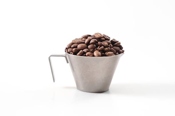 Minimalist image of roasted coffee beans in a small metallic cup with a handle, isolated on a white background. The beans are piled high in the cup, creating a contrast between the dark coffee beans and the bright backdrop.