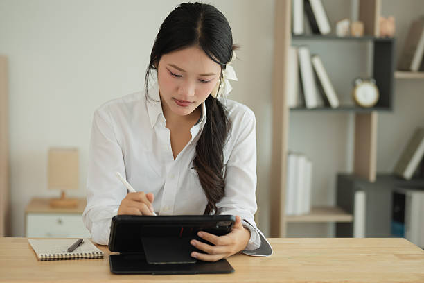 istock Young woman using tablet device to take notes while working at a wooden desk in modern home office with stylish decor and soft lighting 2217572311