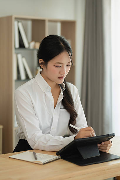 Young Asian woman writing notes on tablet in modern workspace, focused on task, wearing white shirt, reflecting serious and professional demeanor