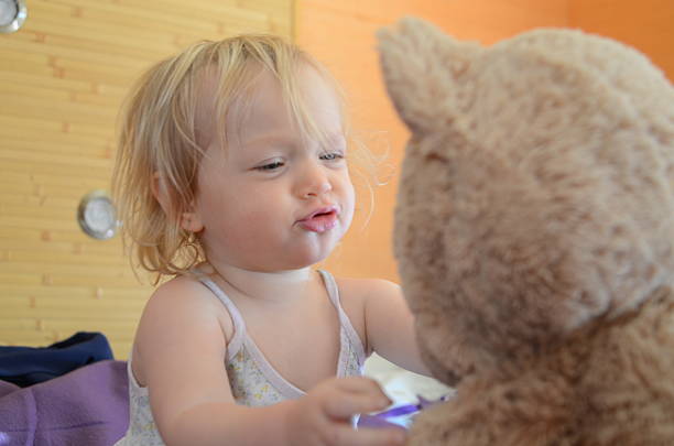 A young child is joyfully interacting with a teddy bear, making playful expressions. The scene captures a moment of innocence and imagination in a cozy indoor setting