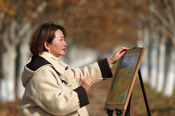 Young Asian woman standing in autumn park with easel, painting oil art and looking at camera. Calm creative lifestyle scene with lake and bridge in the background during cool fall season.