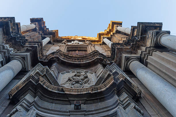 A low angle view of an ornate Baroque facade with intricate stone carvings and columns under a clear blue sky in Italy.