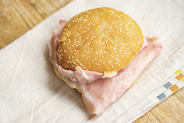 Ham sandwich with thinly sliced cooked ham in a sesame seed bun, on a striped napkin, resting on a rustic wooden table. Ideal for light meals, snacks, or homemade food concepts