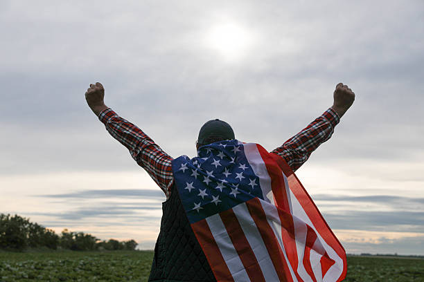 Portrait of cheerful young man looking forward while standing proudly with holding USA flag over his head against wind and blue sky, happy american holding the United states flag celebration